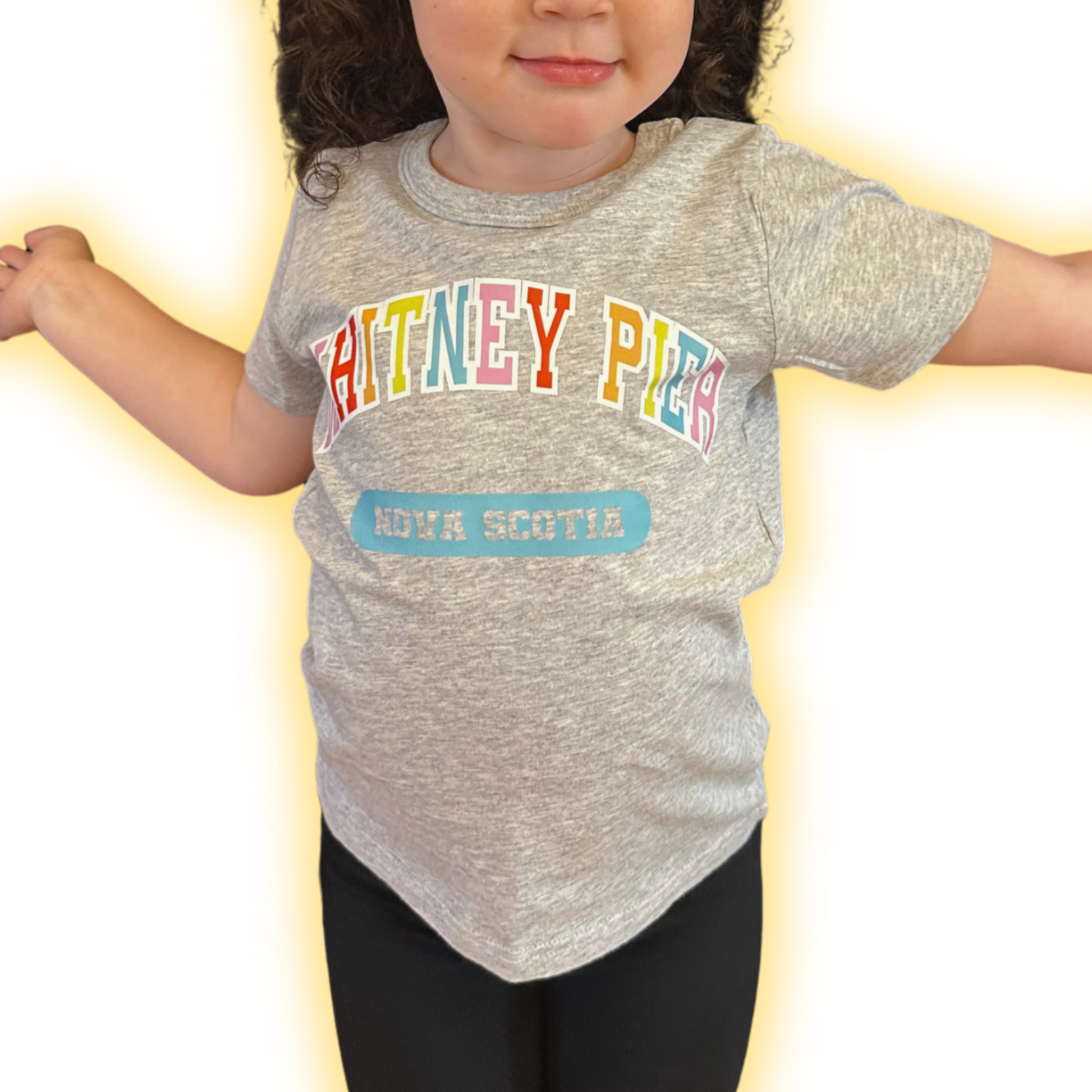 A toddler wearing a heather grey short-sleeve t-shirt with 'WHITNEY PIER' in colourful letters and a blue 'NOVA SCOTIA' patch graphic, paired with black leggings, set against a bright white background with a yellow glow effect.