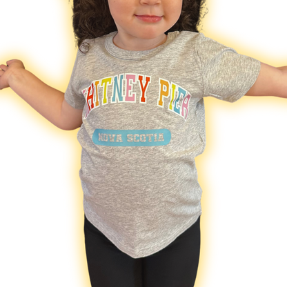 A toddler wearing a heather grey short-sleeve t-shirt with 'WHITNEY PIER' in colourful letters and a blue 'NOVA SCOTIA' patch graphic, paired with black leggings, set against a bright white background with a yellow glow effect.