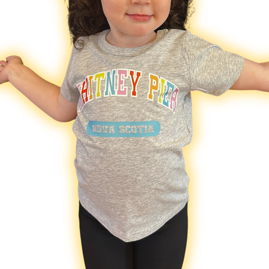 A toddler wearing a heather grey short-sleeve t-shirt with 'WHITNEY PIER' in colourful letters and a blue 'NOVA SCOTIA' patch graphic, paired with black leggings, set against a bright white background with a yellow glow effect.